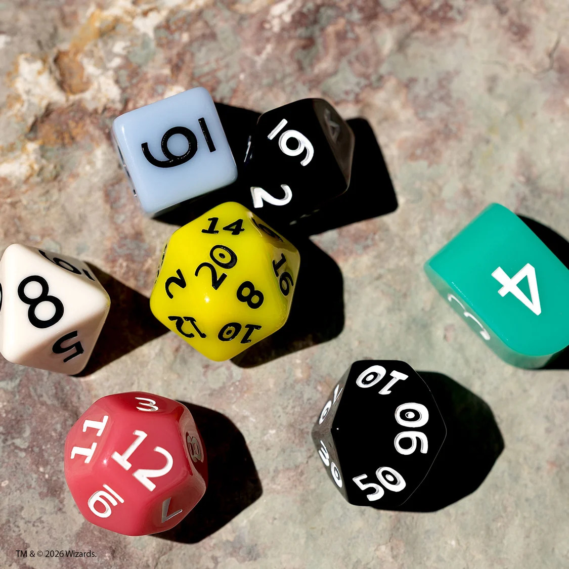 Set of seven polyhedral dice in the colors of the disability pride flag on a stone background.