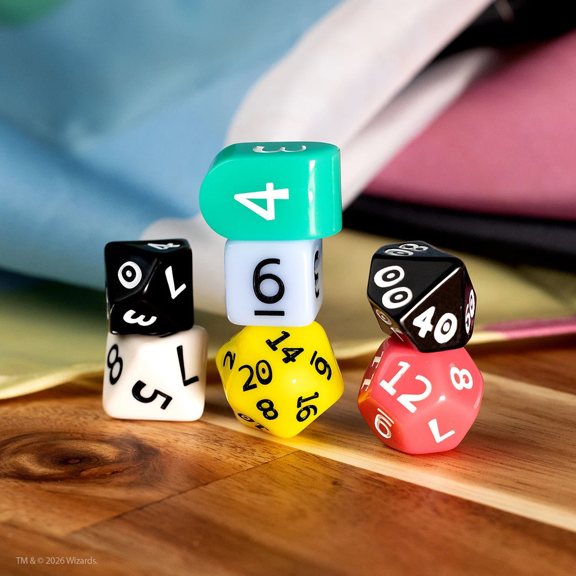 Set of 7 polyhedral dice stacked in three groups. Each dice features a different color from the disability pride flag that sits folded in the background.